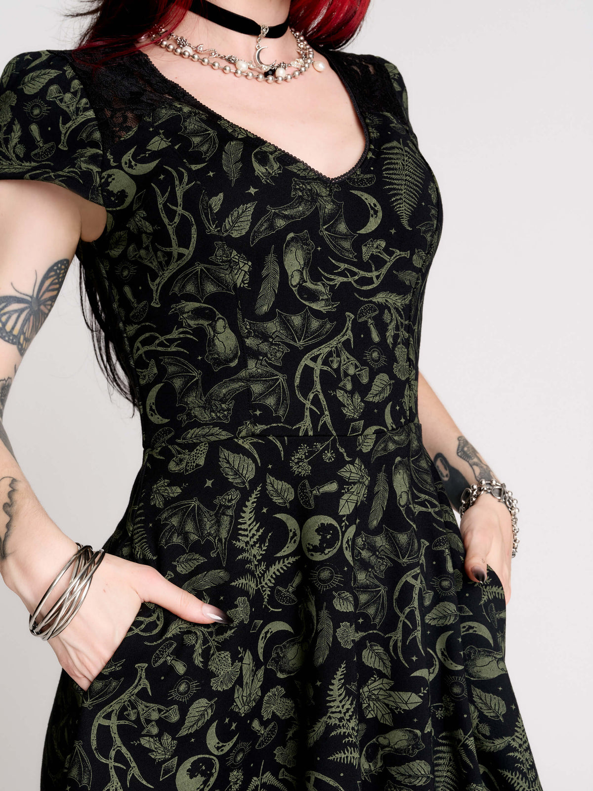 short black dress with a green leafy lace pattern, featuring cap sleeves, a V-neckline, side pockets, and a scalloped lace hem.