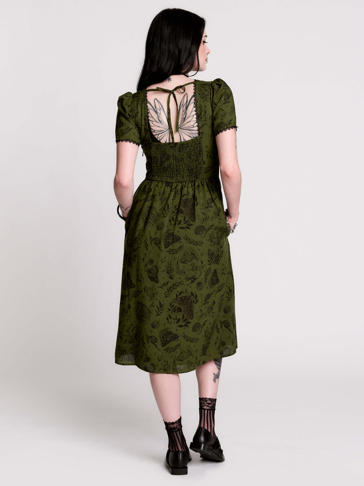 A woman viewed from the back wearing an olive green midi dress with a smocked bodice, short puff sleeves, and a tie-back neckline. The dress has a dark botanical pattern.