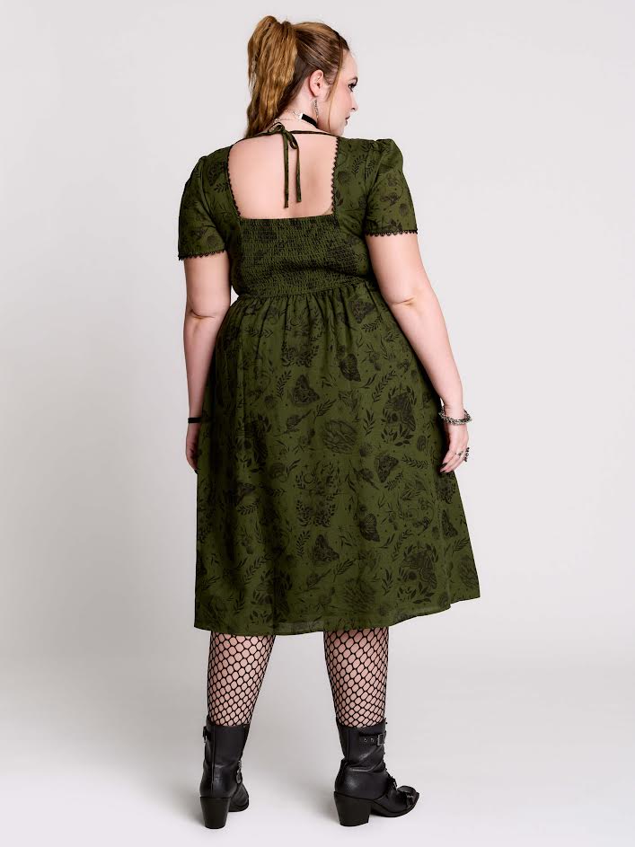 olive green short-sleeve midi dress with a dark floral jacquard pattern, a smocked bodice, and tie-back straps, paired with black fishnet tights and black ankle boots against a light gray studio background.