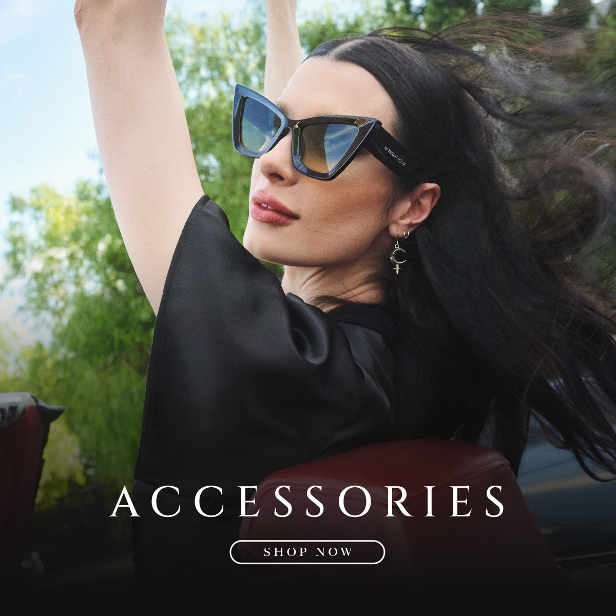 A woman outdoors wearing black cat-eye sunglasses with blue mirrored lenses and a black satin top, with the text 'ACCESSORIES' and 'SHOP NOW' overlaid at the bottom.