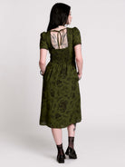A woman viewed from the back wearing an olive green midi dress with a smocked bodice, short puff sleeves, and a tie-back neckline. The dress has a dark botanical pattern.
